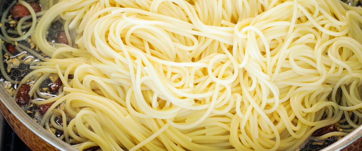 Step 4: Combine Pasta and Pancetta for Cannabis Carbonara Tossing drained spaghetti with crispy pancetta and infused butter in a pan, fourth step for making cannabis-infused carbonara